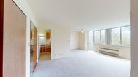 This single-floor residential unit offers a clean, functional layout designed to maximize both space and natural light. The living area is generously sized, finished with neutral wall-to-wall carpeting and bright cream-toned walls that create a light, airy atmosphere. A standout feature is the expansive corner window arrangement, which frames sweeping views of surrounding tree canopies and floods the room with natural light. A ceiling fan adds practicality to the living space, while a baseboard heating unit and a wall-mounted air conditioner ensure year-round comfort.

The kitchen is situated in an open, semi-partitioned layout with a breakfast bar-style counter that connects it to the living area. It is equipped with warm wood-toned cabinetry, a coil-burner electric range with overhead ventilation, white appliances including a refrigerator, and ample counter and storage space. Tile flooring in the kitchen provides a clean contrast to the carpeted living areas.

The bedroom is a well-proportioned, carpet-floored room with a closet accessible through warm wood-toned doors. Additional storage is provided by a built-in shelved closet visible from the main living area. The unit's overall design is practical and understated, offering a neutral canvas well-suited for a variety of interior styles.