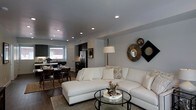 This two-floor home showcases a thoughtfully designed open-concept main level that integrates the living, dining, and kitchen areas into one cohesive, flowing space. The living room is anchored by a generously sized white sectional sofa paired with a sleek round glass coffee table, creating an inviting and contemporary focal point. Recessed lighting throughout the ceiling provides consistent, well-distributed illumination, complemented by an elegant floor lamp that adds warmth to the space. The walls are finished in a soft neutral gray tone, accented by a curated decorative wall arrangement featuring a starburst mirror and geometric décor pieces that add visual interest and personality.

Dark hardwood flooring runs throughout the main level, providing a rich contrast to the light furnishings and walls. The dining area, positioned adjacent to the living space, features a dark wood table and chairs, contributing to the cohesive design language. The kitchen, visible in the background, is equipped with dark cabinetry, stainless steel appliances including a full-sized refrigerator, and benefits from natural light through a window above the sink. The overall ambiance is modern, polished, and welcoming.