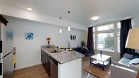 This single-floor space showcases a thoughtfully designed open-concept layout that seamlessly connects the kitchen and living areas. The kitchen is appointed with granite countertops featuring a striking veined pattern, dark espresso cabinetry, a stainless steel undermount sink, and a central island that doubles as a breakfast bar. Three sleek pendant lights hang above the island, providing focused task lighting while contributing to the modern aesthetic.

The kitchen flows naturally into the living area, which is bathed in natural light streaming through large floor-to-ceiling windows dressed with dark curtains. These expansive windows frame views of a lush outdoor landscape and what appears to be a waterway, creating a strong connection to the surrounding environment. The living space features warm hardwood-style flooring that extends throughout, tying both areas together cohesively.

The neutral light grey walls and white ceilings enhance the sense of openness throughout the space. Recessed and flush-mount ceiling fixtures supplement the natural light, ensuring the interior remains well-lit. Overall, the space combines functional design with contemporary finishes to create a bright, inviting atmosphere.