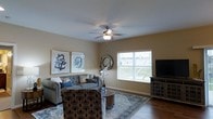 This single-floor residence offers a thoughtfully designed open-concept layout that seamlessly connects the living, dining, and kitchen areas into one cohesive, flowing space. Rich hardwood flooring runs throughout, lending warmth and continuity to the interior.

The kitchen is outfitted with rich cherry-toned cabinetry, granite countertops, stainless steel appliances, and a breakfast bar with seating — ideal for casual dining or entertaining. Pendant lighting above the bar adds a stylish focal point, while recessed lighting ensures the workspace is well-illuminated.

The dining area sits at the heart of the open floor plan, anchored by a classic chandelier and framed by large sliding glass doors fitted with vertical blinds that flood the space with natural light and provide access to the outdoors.

The living room is generously proportioned, featuring warm neutral walls, a ceiling fan with integrated lighting, and large windows that invite additional natural light. The space comfortably accommodates a full seating arrangement and offers a relaxed, welcoming ambiance suitable for everyday living.

Together, the connected areas create an airy, functional layout that balances comfort and practicality, making this single-floor space an inviting and versatile home environment.