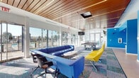 This dynamic single-floor amenity space is designed with a bold, contemporary aesthetic that immediately energizes its occupants. The main lounge area serves as the heart of the facility, featuring generous seating arrangements with vivid blue sectional sofas and striking yellow accent chairs arranged around a central coffee table. A rich wood-slat ceiling with integrated linear lighting runs overhead, adding warmth and architectural depth to the open interior. Floor-to-ceiling glass walls and large sliding doors flood the space with abundant natural light while offering direct views of the outdoor pool area beyond.

A full-size billiards table anchors the entertainment zone, complemented by wall-mounted televisions throughout for added recreation. The colorful patterned carpet ties the lounge and game areas together with a lively, cohesive visual rhythm.

Adjacent to the lounge, a dedicated work and study zone features semi-private pod seating with built-in desktops in red and white, ideal for focused individual use. A glass-walled fitness center is accessible nearby, equipped with treadmills and cardio machines, bathed in natural light from exterior windows.

A bright corridor lined with bold blue accent walls connects the various zones, with a glimpse into a vibrant orange-toned private room, adding yet another layer of color and function to this well-appointed community amenity space.