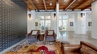 Located at 700 Bloomfield Ave in Bloomfield, Connecticut, this expansive single-floor space offers a thoughtfully designed interior that masterfully blends rustic warmth with contemporary style. Reclaimed wood-plank ceilings with exposed beams run throughout, creating a warm, industrial-chic atmosphere that contrasts beautifully with the polished concrete floors below.

The open layout accommodates multiple functional zones. A central kitchen and bar area features a large island with leather bar stools, pendant globe lighting, dark subway tile backsplash, stainless steel appliances, and dual mounted flat-screen TVs. A dramatic fireplace wall, clad floor-to-ceiling in dark brick with a herringbone firebox surround, anchors a cozy lounge area furnished with leather armchairs and layered vintage rugs.

A generous communal dining area includes a long harvest table beneath a statement pendant light, flanked by open shelving displaying ceramics and pottery. Floor-to-ceiling windows and glass doors along the perimeter flood the space with natural light and offer views of the surrounding greenery.

A separate library lounge features wall-to-wall backlit bookshelves, a media screen, and mixed high-top and lounge seating. Sliding barn doors, frosted glass partitions, and curated artwork complete the eclectic, welcoming character of this distinctive community space.