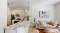 This single-floor residence offers a well-designed open-concept layout that seamlessly connects the kitchen, dining, and living areas. The kitchen is a standout feature, equipped with stainless steel appliances — including an electric range, over-the-range microwave, and refrigerator — complemented by light wood cabinetry with sleek black hardware and granite countertops. A peninsula with seating provides a casual dining option and serves as a natural divider between the kitchen and living space, illuminated by stylish pendant lighting overhead.

The living area is bright and inviting, benefiting from natural light through large windows with blinds. Light wood-tone vinyl plank flooring flows consistently throughout the unit, enhancing the cohesive, contemporary feel.

The bedroom is comfortably sized with ample natural light and connects to an adjacent sitting or flex area, offering additional versatility. The bathroom features a granite-topped vanity with light wood cabinetry, a large mirror, and modern matte black fixtures, mirroring the design language found throughout the unit.

A hallway with clean white paneled doors provides access to the various rooms, maintaining a tidy and organized layout. The overall aesthetic is modern and neutral, with crisp white walls and consistent finishes throughout.