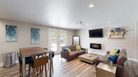 The Village at SoTa's clubhouse serves as the central gathering hub for this Tacoma, WA community. The entrance welcomes visitors with a bright, well-lit foyer showcasing a community map display, decorative wall sconces, and durable wood-look flooring that carries throughout the space. The main lounge area is thoughtfully furnished with plush sofas, a wall-mounted flat-screen TV, a sleek electric fireplace, and floating wood shelving — all accented by contemporary artwork. French doors open directly to the outdoor pool area, filling the lounge with natural light and providing seamless indoor-outdoor connectivity. A dining bar with high-top seating offers an additional casual gathering spot, while a fully equipped kitchen features stainless steel appliances, white shaker cabinetry, a tile backsplash, and stone countertops. A dedicated meeting room provides a more structured setting with a large conference table and white chairs, ideal for community gatherings or events. Outside, a generously sized swimming pool is surrounded by a wide concrete sundeck lined with colorful lounge chairs, all framed by mature evergreen trees. The building exterior is finished in a classic blue, complementing the broader residential community visible in the background.