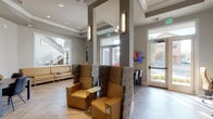 This well-appointed single-floor amenity space combines a welcoming lounge with a fully equipped business center, designed with a cohesive modern aesthetic throughout. The space features striking herringbone-patterned light wood flooring that anchors the entire area, complemented by neutral gray walls, crisp white trim, and tray ceilings with recessed lighting that add architectural dimension.

The lounge area is furnished with plush gold leather seating, including a generous L-shaped sofa and a pair of high-backed accent chairs with decorative upholstery, all accented by slender gold side tables with integrated power outlets. Large interior pass-through windows connect the lounge to adjacent areas, providing visual openness and allowing natural light to flow freely throughout.

The business center features multiple dedicated workstations with dark desks and ergonomic gold leather task chairs, lined along one wall beneath framed artwork. A wall-mounted television and additional seating make this zone equally suited for casual collaboration or focused individual work.

A private meeting or conference area accommodates a small table with modern black chairs, offering a quieter setting for discussions. Elegant wall sconces and a sculptural multi-arm chandelier add warmth and sophistication. Glass entry doors frame views of the outdoor surroundings, reinforcing the bright and open atmosphere throughout the space.