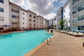 This outdoor amenity space is defined by its expansive resort-style pool area, thoughtfully designed to provide a full range of leisure and relaxation options. The centerpiece is a large, sparkling swimming pool with a separate shallow wading area, both finished with clean tile surrounds and accessible via built-in steps and handrails. The generous pool deck is paved in neutral stone-toned concrete and offers ample room for lounging, with rows of cushioned chaise lounge chairs equipped with blue headrests, side tables, and additional seating clusters shaded by large umbrellas. Elegant white fabric cabanas provide a more private retreat, furnished with plush outdoor sofas and coffee tables. A built-in stone outdoor kitchen and grill station extends the space's entertaining potential. Manicured planters with ornamental trees and shrubs are positioned throughout, adding greenery and visual interest to the hardscape. The entire area is enclosed by sleek black iron fencing, with gated walkways providing controlled access. The surrounding multi-story building features balconies overlooking the courtyard, creating an intimate, community-focused atmosphere. The overall design blends modern architectural elements with comfortable, resort-inspired outdoor living.