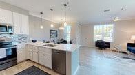 This single-floor residence is thoughtfully designed with a modern, open-concept layout that seamlessly connects the kitchen, dining, and living areas. The kitchen is a standout feature, showcasing white shaker-style cabinetry, quartz countertops, stainless steel appliances, a mosaic tile backsplash, and a central island with pendant lighting overhead. The adjacent dining area offers ample space for a full dining set, while the open living room is bright and airy, featuring crown molding, light wood-look tile flooring, a ceiling fan, and large windows that flood the space with natural light. Sliding glass doors in the living area open to an outdoor view of lush greenery. The two bedrooms are carpeted and well-sized, each accommodating a queen bed comfortably, with generous windows providing pleasant natural light. The primary bathroom features a glass-enclosed walk-in shower with decorative mosaic tile accents and a white vanity with quartz countertops. The second bathroom offers a full tub-shower combination with matching tile detailing and a bright window. Throughout the residence, neutral tones, crown molding, and consistent finishes create a cohesive and polished aesthetic.