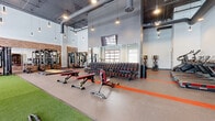 Located at 2870 Pearson Parkway in Plainfield, Indiana, this fitness facility offers a comprehensive workout environment across two distinct exercise rooms. The main exercise area impresses with soaring ceilings, exposed ductwork, and an industrial-modern aesthetic enhanced by pendant lighting and large windows that invite abundant natural light. Signature orange stripe accents on the flooring add a bold visual element throughout the space. This primary room is thoughtfully organized into functional zones: a turf training area ideal for functional and sport-specific drills, a free weight section lined with an extensive dumbbell rack, multiple adjustable benches, strength machines, cable systems, and a row of treadmills and stair climbers. Full-length mirrors along the walls amplify the open, airy feel. A secondary exercise room offers a quieter, more focused environment with light wood-look flooring, clean white walls, and tall ceilings. This room houses interactive cycling bikes, a connected mirror fitness system, and additional cardio equipment, all accessible through sleek glass-panel doors. Together, the two rooms provide a well-rounded fitness experience suited for strength training, cardio conditioning, and functional movement, making this a fully equipped destination for users of all fitness levels.