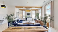 Located at Windsor Hill Blvd in North Charleston, South Carolina, this thoughtfully designed community living room offers a welcoming and well-appointed gathering space. The room makes an immediate impression with its soaring vaulted ceilings, accented by warm exposed wood beams that add architectural character and a touch of coastal charm.

White shiplap walls serve as a distinctive design element, housing a mounted television and built-in cabinetry, while large windows dressed in neutral linen curtains flood the space with natural light and offer views of the surrounding community. The light-toned large-format tile flooring, laid in a herringbone-style pattern, enhances the airy, open feel throughout.

The living area is furnished with a mix of navy blue sofas and accent chairs, warm leather seating, and a live-edge wood coffee table, creating an inviting coastal aesthetic. Statement pendant lights — including a large dome fixture with a gold interior — add a refined touch to the overall design. A white subway tile accent wall features a dual water fountain, reflecting the space's practical community-oriented function. Large-scale abstract artwork further elevates the atmosphere, making this a comfortable and visually engaging space for residents to relax and connect.
