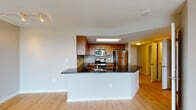 This well-appointed single-floor residence offers a thoughtfully designed open-concept layout that seamlessly connects the main living areas. The kitchen serves as a central highlight, showcasing warm wood cabinetry, dark granite countertops, a mosaic tile backsplash, and a full suite of stainless steel appliances including a range, microwave, and refrigerator. A skylight above the kitchen floods the space with natural light, while a wraparound peninsula island provides additional prep space and a natural division between the kitchen and the living area. Light hardwood-style flooring runs throughout the main living spaces, contributing to a bright and cohesive aesthetic. The spacious living area features floor-to-ceiling sliding glass doors that open to a balcony and allow for abundant natural light and outdoor views. The bedroom is generously sized, featuring plush carpeting, large windows with vertical blinds, and direct balcony access. The bathrooms are finished with matching dark granite countertops, wood-toned cabinetry, and light tile flooring. The primary bathroom offers a double-sink vanity with modern fixtures, while the secondary bathroom includes a full soaking tub and shower combination. Throughout the space, the design maintains a clean, contemporary aesthetic with neutral tones and quality finishes.