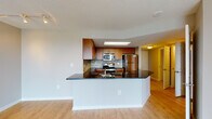 This well-appointed single-floor residence offers a thoughtfully designed open-concept layout that seamlessly connects the main living areas. The kitchen serves as a central highlight, showcasing warm wood cabinetry, dark granite countertops, a mosaic tile backsplash, and a full suite of stainless steel appliances including a range, microwave, and refrigerator. A skylight above the kitchen floods the space with natural light, while a wraparound peninsula island provides additional prep space and a natural division between the kitchen and the living area. Light hardwood-style flooring runs throughout the main living spaces, contributing to a bright and cohesive aesthetic. The spacious living area features floor-to-ceiling sliding glass doors that open to a balcony and allow for abundant natural light and outdoor views. The bedroom is generously sized, featuring plush carpeting, large windows with vertical blinds, and direct balcony access. The bathrooms are finished with matching dark granite countertops, wood-toned cabinetry, and light tile flooring. The primary bathroom offers a double-sink vanity with modern fixtures, while the secondary bathroom includes a full soaking tub and shower combination. Throughout the space, the design maintains a clean, contemporary aesthetic with neutral tones and quality finishes.