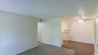 This single-floor space presents a functional and straightforward layout with a notable mix of materials and finishes throughout. The living area is finished with wall-to-wall carpet, providing a comfortable underfoot feel, while the adjoining kitchen area transitions to hard flooring. A standout architectural detail is the diagonal wood-paneled accent wall, painted white, which adds a subtle textural element to the space. The textured popcorn ceiling runs throughout, and a central wall partition separates the main living zone from the kitchen area, creating a semi-defined layout without fully closing off either space. The kitchen features white upper and lower cabinetry with ample storage, offering a clean, neutral aesthetic. A louvered closet door is visible near the partition, indicating additional storage options within the space. An HVAC vent is positioned on the upper wall, ensuring climate control throughout. The overall color palette is neutral, with white walls, light ceilings, and understated finishes, creating a blank canvas that is move-in ready and adaptable to a variety of interior styles.