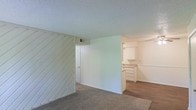 This single-floor space presents a functional and straightforward layout with a notable mix of materials and finishes throughout. The living area is finished with wall-to-wall carpet, providing a comfortable underfoot feel, while the adjoining kitchen area transitions to hard flooring. A standout architectural detail is the diagonal wood-paneled accent wall, painted white, which adds a subtle textural element to the space. The textured popcorn ceiling runs throughout, and a central wall partition separates the main living zone from the kitchen area, creating a semi-defined layout without fully closing off either space. The kitchen features white upper and lower cabinetry with ample storage, offering a clean, neutral aesthetic. A louvered closet door is visible near the partition, indicating additional storage options within the space. An HVAC vent is positioned on the upper wall, ensuring climate control throughout. The overall color palette is neutral, with white walls, light ceilings, and understated finishes, creating a blank canvas that is move-in ready and adaptable to a variety of interior styles.