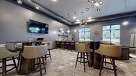 Located at 6620 Chestnut St in Lino Lakes, MN, this single-floor dining and lounge space offers a sophisticated, contemporary design with a well-appointed layout. The room features soaring ceilings with exposed ductwork that adds an industrial-chic character, complemented by cool grey walls that create a sleek, modern atmosphere throughout.

Lighting is a standout element, with striking sputnik-style chandelier fixtures and globe pendant lights suspended at varying heights, casting a warm, ambient glow across the space. The patterned, tile-effect carpet flooring adds texture and visual interest underfoot.

The room is divided into distinct functional zones. A wet bar area along one wall includes a mosaic tile backsplash, open shelving, a stainless steel refrigerator, and a counter lined with swivel bar stools — ideal for casual dining or socializing. Multiple high-top tables with padded barrel chairs are arranged throughout, providing ample communal seating. A dedicated lounge corner features plush upholstered armchairs and a high-backed booth-style seat, offering a more relaxed setting.

Large windows along one wall flood the space with natural light and offer views of the surrounding greenery. A wall-mounted flat-screen TV and vibrant artwork displayed on the walls round out this well-designed, multifunctional gathering space.