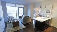 This single-floor residence showcases a thoughtfully designed open-concept layout that connects the kitchen and living areas into one cohesive, functional space. Dark hardwood flooring runs throughout, grounding the interior with a rich, contemporary aesthetic that contrasts beautifully against the light-toned walls and bright white surfaces.

The living area is anchored by a comfortable seating arrangement positioned atop a plush area rug, with floor-to-ceiling windows and a glass-paneled balcony door flooding the space with abundant natural light. The expansive glazing frames striking urban skyline views, giving the room an airy, elevated feel.

The kitchen features a sleek island with a crisp white countertop and an integrated sink, complemented by dark cabinetry that echoes the tones of the flooring. A modern pendant light hangs above, adding a refined focal point to the culinary space.

A corridor visible from the main living area leads to additional rooms, including a bedroom appointed with warm lighting and woven accent furnishings, maintaining the space's cohesive modern sensibility throughout. Overall, the residence offers a polished, urban living experience.