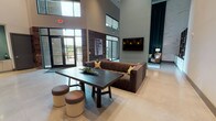The clubhouse at Millennium Place in Corinth, Texas, is a thoughtfully designed single-floor community space that makes a strong architectural impression through its soaring double-height ceilings and expansive, open layout. Large floor-to-ceiling windows and glass entry doors flood the interior with natural light while offering views of the surrounding outdoor amenity areas, including a glimpse of the pool beyond the main entrance.

The main lounge area anchors the space with a generous sectional sofa, organic wood accent tables, and a wall-mounted flat-screen television. A reclaimed wood feature wall adds warmth and texture, contrasting beautifully with the polished concrete floors and crisp white walls. Curated artwork throughout contributes to the space's gallery-like quality.

A dedicated seating nook offers a more intimate atmosphere, furnished with a teal sofa, a leather wingback chair, and floor-length emerald drapery that frames a dramatic marble-tiled accent wall. Adjacent to the main lounge, a bar-height counter with a textured stone backsplash and amber pendant lighting provides casual seating and a social gathering point. The overall ambiance is contemporary and welcoming, balancing bold design elements with comfortable, functional furnishings.