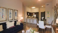 This single-floor space showcases an open-concept layout that seamlessly integrates the living, dining, and kitchen areas into one cohesive environment. The living area is anchored by a deep navy sofa accented with neutral throw pillows, complemented by a sleek glass coffee table and warm ambient lighting from a classic table lamp. Framed artwork adorns the walls, adding a curated aesthetic to the space. Adjacent to the living area, a dining space features a dark-toned table paired with white upholstered chairs, creating a refined contrast. The kitchen is a standout feature, boasting dark cabinetry, stainless steel appliances, and a breakfast bar with bar-height seating illuminated by pendant lighting. The light-toned countertop surface adds brightness against the rich cabinetry. Throughout the space, wood-look flooring provides warmth and continuity. The tall, smooth walls and recessed lighting contribute to a sense of openness and height. The overall ambiance is modern and sophisticated, with a harmonious blend of dark and light tones that creates an inviting yet stylish atmosphere.