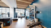 This fitness center at 2703 Girard Ave S in Minneapolis, Minnesota, is a thoughtfully designed workout space that maximizes every square foot to deliver a comprehensive exercise experience. The room is defined by a striking deep teal accent wall adorned with motivational typography, creating an energizing ambiance that sets the tone for an effective workout.

The ceiling features a distinctive slatted wood design with integrated linear LED lighting, lending the space a modern, architectural character. Large street-facing windows along the front wall flood the interior with natural light and offer views of the surrounding Lyndale neighborhood, enhancing the open feel of the space.

Durable rubber flooring in a warm tan tone runs throughout, providing a practical and resilient surface suited for a variety of exercises. Equipment is efficiently arranged to make the most of the layout. Cardio offerings include treadmills, an elliptical, and a rowing machine positioned near the windows. A dual cable cross machine and a full dumbbell rack with an adjustable bench support strength training needs. A wall-mounted rack stores kettlebells, medicine balls, and exercise mats, while plyometric boxes add functional training options. The space is accessed through sleek glass-panel doors, contributing to its clean, contemporary look.