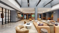 Located at 316 15th St in Jersey City, NJ, this striking single-floor lobby space spans an expansive footprint, offering a sophisticated blend of industrial character and modern design. Polished concrete floors flow seamlessly throughout, reflecting natural light that pours in through dramatic floor-to-ceiling steel-framed windows along the street-facing façade — a defining architectural feature that floods the interior with an open, airy ambiance.

The main living and lounge area is anchored by soaring ceilings with skylights, exposed structural columns, and textured exposed brick walls that add warmth and historic character to the contemporary setting. Multiple seating arrangements — featuring tan leather sofas, curved accent chairs, and marble-top coffee tables — are organized to create distinct yet open social zones. Curated contemporary artwork adorns the walls throughout.

A dedicated hallway corridor connects the lounge to a staffed reception desk, accented by geometric pendant lighting and dark charcoal wall finishes. A separate, glass-partitioned dining or meeting nook offers additional flexible space. Decorative indoor greenery adds life and softness to the otherwise industrial palette. The entrance vestibule features bold black-framed glass doors, offering clear sightlines to the surrounding Jersey City streetscape.