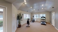 This community fitness center offers a dedicated space for cardiovascular exercise within a single-floor layout designed for functionality and comfort. The space features vaulted ceilings accented by recessed lighting, which contribute to an open and airy atmosphere throughout. Neutral-toned walls and wall-to-wall carpeting create a clean, approachable environment suited for regular workouts.

The main exercise area is outfitted with a selection of cardio equipment, including a treadmill, an elliptical trainer, and a recumbent stationary bike, providing users with a variety of workout options. Large windows line multiple walls, inviting abundant natural light and offering views of the surrounding community, including glimpses of a pool area and adjacent buildings.

A separate entry zone near the front door features tile flooring and a small cabinetry unit, providing a practical transition space. The room also includes a security camera and posted community guidelines, reflecting a well-managed and safety-conscious facility. A decorative potted palm plant adds a touch of greenery to the otherwise utilitarian space. Multiple access doors and ventilation fixtures throughout further support the comfort and usability of this community amenity.