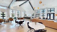 Located at 5617 Oakley Industrial Blvd in South Fulton, GA, this expansive single-floor amenity space is a masterclass in modern interior design. Soaring vaulted ceilings with bold exposed dark beams define the architectural character, while floor-to-ceiling black-framed windows flood the interior with natural light and offer clear views of the surrounding community.

The open-plan layout is thoughtfully organized into distinct yet connected zones. A lounge area anchors one end of the space, centered around a sleek linear fireplace set against a dramatic textured accent wall with complementary botanical wallpaper. Comfortable sectional seating, a large-screen TV, and curated pendant lighting create an inviting atmosphere.

A dedicated game area features foosball and billiard tables alongside casual seating clusters, while a dining and co-working zone is highlighted by a striking living green wall column and warm wood-slat ceiling details. Round café-style tables with modern black chairs are distributed throughout for flexible use.

The kitchenette area is fully outfitted with marble-topped counters, open shelving, a white subway tile backsplash, and brass fixtures, bathed in light from tall windows. Polished concrete floors run seamlessly throughout, grounding the space with an industrial-chic aesthetic. Dual entry doors open directly to an outdoor terrace, extending the social experience beyond the interior.