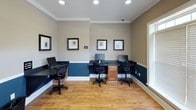 This single-floor business center is designed to provide a functional and professional workspace. The room features a distinctive two-tone wall treatment, with deep navy blue on the lower half and warm beige on the upper half, separated by crisp white chair rail molding. Crown molding adds a refined architectural detail along the ceiling line.

The workstation area accommodates multiple users simultaneously, with dark-surfaced desks lining the walls and center of the room, each equipped with desktop computers, keyboards, and ergonomic task chairs. A warm-toned wood cabinet provides built-in storage beneath the central desk configuration. Framed botanical artwork decorates the walls, contributing to a polished, professional atmosphere.

Natural light filters into the space through large windows fitted with horizontal blinds, including transom windows above that allow additional daylight. Warm hardwood-style flooring runs throughout, complementing the overall color palette. A printer or copier station is positioned near the windows for added convenience. Recessed lighting and a glass-paneled door complete the space, ensuring both adequate illumination and easy access. Signage indicates a no food or drinks policy, maintaining the integrity of the equipment.
