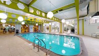 This single-level indoor aquatic and fitness facility is defined by its vibrant design and well-appointed amenities. The centerpiece is a generously sized indoor lap pool with crystal-clear turquoise water, flanked by a broad tiled pool deck furnished with contemporary lounge chairs. Oversized spherical pendant lights hang from the exposed ceiling structure, which features bold lime-green beams and hexagonal cutouts that give the space a modern, energetic character.

Natural light pours in through floor-to-ceiling windows and glass doors positioned along multiple walls, offering views of surrounding outdoor terraces and greenery. These glass panels create a seamless visual connection between the interior and exterior, enhancing the open, airy atmosphere throughout.

A circular hot tub is tucked into a dedicated alcove finished in white brick tile, complete with safety equipment mounted on the walls. Adjacent to the pool area, a well-equipped fitness center is visible through glass partitions, housing cardio and strength-training machines under equally bright, high ceilings.

The pool surround transitions into a relaxation zone with additional seating and direct access to an outdoor terrace. Pale stone tile flooring runs throughout, tying together the various areas of the facility into a cohesive, resort-like environment.