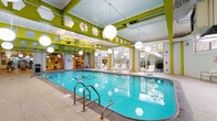 This single-level indoor aquatic and fitness facility is defined by its vibrant design and well-appointed amenities. The centerpiece is a generously sized indoor lap pool with crystal-clear turquoise water, flanked by a broad tiled pool deck furnished with contemporary lounge chairs. Oversized spherical pendant lights hang from the exposed ceiling structure, which features bold lime-green beams and hexagonal cutouts that give the space a modern, energetic character.

Natural light pours in through floor-to-ceiling windows and glass doors positioned along multiple walls, offering views of surrounding outdoor terraces and greenery. These glass panels create a seamless visual connection between the interior and exterior, enhancing the open, airy atmosphere throughout.

A circular hot tub is tucked into a dedicated alcove finished in white brick tile, complete with safety equipment mounted on the walls. Adjacent to the pool area, a well-equipped fitness center is visible through glass partitions, housing cardio and strength-training machines under equally bright, high ceilings.

The pool surround transitions into a relaxation zone with additional seating and direct access to an outdoor terrace. Pale stone tile flooring runs throughout, tying together the various areas of the facility into a cohesive, resort-like environment.