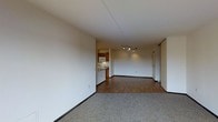 This single-floor space offers an open-concept layout that seamlessly connects the living area to the kitchen. The living space features neutral-toned carpet, providing a comfortable and inviting foundation, while the remainder of the unit transitions to hardwood-style flooring that extends toward the kitchen and beyond. The walls are finished in a clean, off-white tone throughout, contributing to a bright and airy atmosphere.

A track lighting fixture anchors the main living and dining area, offering adjustable, functional illumination across the open space. The compact kitchen is positioned toward the rear of the unit and is equipped with warm wood-toned cabinetry on both upper and lower sections, a built-in microwave, and a range with a ventilation hood. White lower cabinetry adds contrast beneath the counter space, keeping the kitchen feel clean and organized.

A doorway visible along the far wall suggests access to additional rooms, likely a bedroom or utility area. Dark baseboards throughout provide a subtle but defined contrast against the light walls and flooring, adding a polished finishing touch to the overall interior.