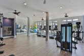 Located at 83 Ernest W Barrett Pkwy NW in Marietta, Georgia, this well-appointed fitness center offers a comprehensive workout environment designed for both functionality and comfort. The single-floor facility features warm wood-look flooring throughout, white structural columns, and ceiling fans that contribute to a bright, airy atmosphere.

The cardio zone is a standout feature, lined with floor-to-ceiling windows that flood the space with natural light and offer direct views of the outdoor pool area and surrounding greenery. Multiple treadmills, upright stationary bikes, and elliptical machines are positioned to take full advantage of these scenic outdoor views.

A dedicated strength training area anchors one side of the facility, featuring a multi-functional cable machine, weight bench, and strength equipment. Bold blue accent walls define this zone and add a dynamic contrast to the clean white trim and light flooring. A wall-mounted television provides entertainment during workouts.

An alcove area, highlighted with deep purple accent walls, houses a water station with cabinetry, providing convenient hydration access. Adjacent to this, a well-stocked rack of colorful kettlebells and a full-length mirror support free weight and functional training workouts. A separate area accommodates stability balls and resistance accessories, rounding out the facility's versatile offerings. The overall ambiance is energetic yet inviting, catering to a wide range of fitness needs.