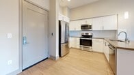 Located at 770 S Harbor Blvd in Fullerton, CA, this well-designed two-bedroom, two-bathroom residence offers a thoughtful single-floor layout with a clean, contemporary aesthetic. The open-concept kitchen and dining area serves as the heart of the home, showcasing white shaker cabinetry, quartz-style countertops, a peninsula with pendant lighting, and a full suite of stainless steel appliances including a refrigerator, range, and microwave. Light wood-tone flooring runs throughout the main living areas, complementing the neutral color palette and recessed lighting overhead. The living and dining space flows naturally toward a private balcony, where large windows invite abundant natural light and outdoor views of the surrounding greenery. Both bedrooms feature plush carpeting and generous closet space with built-in shelving and hanging rods — three closets total ensure ample storage throughout the unit. The primary bedroom includes its own en-suite bathroom, appointed with a wide vanity, white cabinetry, a taupe countertop, and an illuminated mirror. The second bathroom features a full tub-shower combo, matching white cabinetry, and light tile flooring. A hallway connects all spaces efficiently, completing this well-appointed and move-in-ready home.
