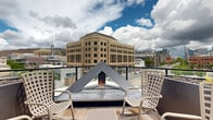 This thoughtfully designed rooftop amenity deck offers a versatile outdoor retreat set against a striking urban backdrop. The space is organized into distinct functional zones, each finished with high-quality materials including large-format stone pavers and warm wood-toned decking that add texture and visual interest throughout.

At the heart of the deck, a lush artificial turf lawn provides a vibrant green centerpiece, bordered by raised Corten steel planting beds with seasonal greenery. Colorful modular seating punctuates the lawn area, creating casual gathering spots. String lights are strung overhead, lending a warm, festive ambiance that extends into the evening hours.

An outdoor kitchen station features dual stainless steel grills, prep counters, and a sink — ideal for al fresco dining and entertaining. Multiple seating arrangements are distributed across the deck, including dining tables with chairs and bench-style seating with wood accents, accommodating both intimate gatherings and larger groups.

A striking angular fire feature serves as a focal point on the perimeter terrace, flanked by lounge chairs and framed by panoramic city and mountain views. An outdoor television and dining area offer additional entertainment options.

The surrounding multi-story building facade, clad in contemporary metal paneling and warm wood accents, encloses portions of the deck while keeping it open to the sky — filling the space with natural light and fresh air throughout the day.
