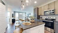 This well-designed one-bedroom, one-bathroom unit offers a thoughtfully planned single-level layout that maximizes every square foot. The open-concept kitchen and living area serves as the heart of the home, anchored by a substantial kitchen island with dark countertops and seating for multiple guests, illuminated by globe pendant lights. The kitchen features a two-tone cabinet design combining gray shaker-style lower cabinets with warm wood-toned uppers, a subway tile backsplash, and a full suite of stainless steel appliances. An in-unit washer and dryer is conveniently situated adjacent to the kitchen area. The living room is bright and airy, benefiting from natural light streaming through large windows and a glass-paneled door that opens onto a private balcony with outdoor views. Wood-style flooring runs throughout the main living areas, adding warmth and continuity. The bedroom is a calm retreat with plush carpet, a dedicated window for natural light, and direct access to a walk-in closet equipped with built-in shelving. The bathroom features gray cabinetry, a white countertop, a large mirror, and a glass-enclosed shower with tile surround.