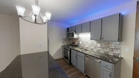 This single-floor unit offers a thoughtfully updated interior with a clear distinction between its living, sleeping, and cooking spaces. The kitchen is the standout feature, showcasing grey shaker-style cabinetry with brushed nickel hardware, dark granite countertops, and a full suite of stainless steel appliances including a gas range, built-in microwave, refrigerator, and dishwasher. A mosaic tile backsplash in neutral tones of beige, brown, and silver adds texture and visual interest, while ambient blue LED lighting along the upper cabinets creates a modern atmosphere. A peninsula with matching countertop extends into the adjoining living area, which is illuminated by a chandelier overhead. The living space transitions from hardwood-style flooring in the kitchen area to plush carpeting, and large windows with vertical blinds allow natural light to fill the room generously. The bedroom is spacious and carpeted, featuring a wall of sliding mirrored closet doors that enhance the sense of openness. The bathroom is clean and functional, with light tile flooring, a shower-tub combination, granite-topped vanity, and simple brushed nickel fixtures throughout.