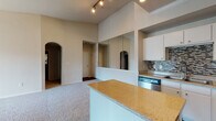 This single-floor residence showcases a thoughtfully designed open-concept layout that seamlessly connects the living and kitchen areas. Soaring vaulted ceilings with track lighting lend an airy, expansive feel to the main living space, which is finished with plush carpeting and accented by a ceiling fan and elegant arched doorways. A large floor-to-ceiling mirror further amplifies the sense of space throughout.

The kitchen is a standout feature, equipped with a full suite of stainless steel appliances including a refrigerator, range, microwave, and dishwasher. White cabinetry pairs cleanly with granite-style countertops and a striking mosaic tile backsplash. A central island provides additional prep and serving space, while rich dark hardwood flooring grounds the room with warmth and contrast.

The primary bathroom offers a long vanity with granite-style countertops, white cabinetry, and direct access to a generously sized walk-in closet outfitted with wire shelving on multiple walls. A secondary bathroom features a tiled tub-shower combo with overhead cabinetry for added storage.

Bedroom spaces are finished with soft carpeting and neutral walls, offering a calm and versatile canvas. Throughout the residence, a consistent neutral color palette and quality finishes create a cohesive and comfortable living environment.