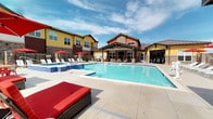 This expansive outdoor amenity area centers around a generously sized swimming pool with crystal-clear blue water, complemented by an adjacent spa with decorative blue tile accents and a water feature. The pool deck is finished in large-format concrete pavers and provides ample space for lounging, furnished with red cushioned chaise loungers, wicker seating, and bold red umbrellas that add a lively splash of color throughout the space.

A covered pavilion anchored by stacked-stone columns offers a shaded retreat with comfortable seating, providing an ideal vantage point overlooking the pool. The combination of natural stone masonry and wood-beam detailing gives the pavilion a warm, craftsman-inspired character. A dedicated pool house structure, clad in rich red-orange lap siding with stone base accents, provides facility access and posted guidelines for guests.

The entire area is enclosed by a wrought-iron fence with stacked-stone pillars topped with wall-mounted light fixtures, lending a refined and secure perimeter to the space. The surrounding multi-story residential buildings feature a vibrant palette of yellow, red, and earth tones with metal rooflines and balconies that frame the courtyard. Open skies above allow abundant natural light to flood the deck, creating a bright and inviting outdoor retreat.