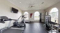 This fitness center offers a clean, functional environment designed to support a comprehensive workout routine. The space is housed on a single floor and features vaulted ceilings accented by a ceiling fan, contributing to an open and airy atmosphere. Durable dark rubber flooring covers the entire area, providing both safety and stability for exercise.

Natural light floods the space through multiple arched windows with elegant fanlights, offering views of the surrounding exterior and creating a bright, welcoming ambiance. Wall-mounted flat-screen televisions are positioned for easy viewing during cardio sessions.

The cardio zone includes multiple treadmills and elliptical trainers from reputable brands, accommodating a range of fitness levels. The strength training area is well-appointed with cable machines, a multi-station weight system, a recumbent bike, and a dedicated workout bench, allowing for a full-body training regimen. A small accessory station with free weights and fitness accessories is also available.

French doors with shuttered windows provide an additional access point and allow for natural ventilation. Colorful artwork adorns the walls, adding a touch of personality to the otherwise clean and neutral aesthetic. The overall layout is thoughtfully organized to maximize usable floor space while keeping equipment accessible and easy to navigate.