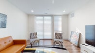 This single-floor residence showcases a thoughtfully designed interior with a clean, contemporary aesthetic throughout. The living area is bright and welcoming, characterized by warm hardwood flooring, recessed lighting, and large windows dressed with layered horizontal sheer blinds that allow diffused natural light to filter in. The seating arrangement includes a cognac leather sofa, bold chevron-patterned accent chairs, a glass coffee table, and a geometric area rug, creating a visually dynamic yet cohesive space.

The kitchen is a standout feature, boasting sleek white cabinetry, striking marble-look surfaces on both the backsplash and peninsula, and stainless steel appliances including a side-by-side refrigerator and built-in microwave. A peninsula counter with white upholstered bar stools provides a casual dining option and defines the kitchen's open connection to the living area.

The bedroom offers a calm, neutral retreat with the same warm hardwood flooring, built-in low-profile white storage, a mirrored closet, and tasteful wall art. The bathroom is elegantly finished with floor-to-ceiling marble-look tile, a soaking tub, and a frameless sliding glass shower enclosure, delivering a spa-like atmosphere with refined, polished details.