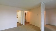 This single-floor space presents a practical and functional layout designed to maximize utility. The main living area features wall-to-wall carpeting in a neutral beige tone, creating a warm and understated foundation. A textured ceiling adds subtle visual character throughout. A louvered bifold door is visible to the right, likely concealing a closet and offering convenient built-in storage.

Two arched openings connect the living area to the adjoining spaces. To the left, the kitchen is equipped with full-height white cabinetry that provides generous storage capacity, complemented by hard flooring that contrasts with the carpeted living zone. The clean, bright cabinetry contributes a fresh, organized appearance to the kitchen area.

The second opening leads to a utility area, where a water heater is visible, indicating practical infrastructure tucked within the unit's footprint. Hard flooring continues into this zone, making maintenance straightforward. A thermostat panel is mounted on the main living area wall, offering convenient climate control access. Overall, the space presents a clean, move-in ready environment with a no-frills, utilitarian character suited for everyday living.