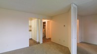 This single-floor space presents a practical and functional layout designed to maximize utility. The main living area features wall-to-wall carpeting in a neutral beige tone, creating a warm and understated foundation. A textured ceiling adds subtle visual character throughout. A louvered bifold door is visible to the right, likely concealing a closet and offering convenient built-in storage.

Two arched openings connect the living area to the adjoining spaces. To the left, the kitchen is equipped with full-height white cabinetry that provides generous storage capacity, complemented by hard flooring that contrasts with the carpeted living zone. The clean, bright cabinetry contributes a fresh, organized appearance to the kitchen area.

The second opening leads to a utility area, where a water heater is visible, indicating practical infrastructure tucked within the unit's footprint. Hard flooring continues into this zone, making maintenance straightforward. A thermostat panel is mounted on the main living area wall, offering convenient climate control access. Overall, the space presents a clean, move-in ready environment with a no-frills, utilitarian character suited for everyday living.