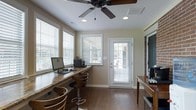 This single-floor space combines functional design with warm, inviting aesthetics. The interior is anchored by rich, dark hardwood flooring that runs throughout, complementing the neutral beige walls and white trim for a cohesive, polished look. A standout feature is the built-in granite-topped countertop workstation, which runs along one wall beneath a bank of windows, offering ample workspace alongside stylish bar-height seating with modern swivel stools. Multiple windows dressed with white horizontal blinds bathe the space in natural light while maintaining privacy. A ceiling fan with a dark finish adds both visual interest and practical airflow management overhead, alongside recessed lighting for well-distributed illumination. A white-framed glass-panel door provides direct access to an outdoor area, further enhancing the sense of openness. An exposed brick accent wall adds a touch of texture and character. A wooden sideboard or media console is also present, adding to the functional storage options. Overall, the space strikes a balance between a comfortable work environment and relaxed living, making it highly versatile for everyday use.