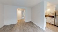 This single-floor residential unit offers a well-organized layout with a clean, contemporary aesthetic throughout. The open living area is bathed in natural light from a large picture window dressed with sheer curtains, highlighting the smooth luxury vinyl plank flooring and neutral-toned walls. A textured ceiling adds subtle character to the space, while a built-in wall unit provides convenient climate control.

The galley-style kitchen is thoughtfully designed with white cabinetry, dark countertops, and stainless steel appliances including a full-size refrigerator, electric range, and dishwasher. Track lighting overhead ensures excellent task illumination, and the same light vinyl plank flooring extends seamlessly throughout.

A hallway connects the living areas to the private bedroom, which features soft wall-to-wall carpeting, a ceiling fan with integrated lighting, and a window overlooking lush greenery — creating a calm and comfortable retreat. The full bathroom is finished in a crisp, all-white palette, complete with a tiled shower-tub combination, brushed nickel fixtures, and coordinating wood-look tile flooring that ties the space together. The overall design is clean, functional, and move-in ready.