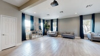 This single-floor space combines functionality with modern design, making it well-suited for gatherings, events, or community use. The main open area is expansive and airy, featuring light wood-look tile flooring that runs consistently throughout the entire space. Recessed lighting illuminates the interior, complemented by abundant natural light streaming through multiple windows dressed with bold navy blue grapnel-style curtains, lending a polished and cohesive aesthetic. A ceiling-mounted projector adds a practical media capability to the main room, which is currently furnished with neutral-toned sofas and accent consoles.

A glass partition wall separates the main area from an adjoining room, offering both visual connectivity and flexible spatial division. The kitchen is a standout feature, boasting navy-toned shaker-style cabinetry, a bold mosaic tile backsplash, granite countertops, a large island with pendant lighting overhead, and a full suite of stainless steel appliances. A vivid navy accent wall adds a dramatic focal point to the kitchen area.

Throughout the space, warm gray walls, clean white trim, and consistent flooring create a unified, contemporary atmosphere that feels both inviting and versatile.
