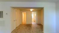 This two-floor residential space offers a functional and straightforward layout designed for comfortable everyday living. The main level features a spacious carpeted living area that opens through a wide archway into an adjoining room, creating a connected and flowing floor plan. A ceiling light fixture provides overhead illumination, complementing the warm, neutral tones found throughout the space. Just beyond the archway, a dedicated kitchen area is visible, outfitted with wooden cabinetry and a window that allows natural light to filter in, brightening the cooking and dining space. The kitchen's tile or vinyl flooring contrasts with the plush carpeting of the living area, clearly defining each functional zone. A hallway accessible from the main living space leads further into the home, connecting to additional rooms on the ground floor. Closet space is present near the entryway, adding convenient storage. The overall color palette consists of soft, neutral creams and beiges, lending the interior a calm and understated ambiance. Spread across two floors, the home provides ample room for varied living arrangements, with the upper level offering additional private spaces. The layout is practical and well-organized, making it a versatile residential space suited for a range of occupants.