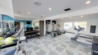 This fitness center offers a comprehensive workout environment designed to accommodate a variety of exercise routines. The space is finished with a distinctive geometric-patterned carpet in soft grey and teal tones, adding a modern, energetic aesthetic to the room. Recessed lighting throughout the ceiling ensures consistent, even illumination across the entire area.

The cardio section is well-appointed with multiple elliptical machines and a treadmill, each paired with a dedicated flat-screen television mounted at eye level for entertainment during workouts. A large window along one wall allows natural light to filter in and provides an outdoor view, enhancing the open feel of the space.

A full rack of dumbbells in graduated weights lines one wall, catering to strength training needs. A large mirror is positioned nearby, allowing users to monitor their form during exercises. An accent wall painted in a bold teal blue adds a vibrant focal point to the room and complements the overall color palette.

The space also features a connected room visible through a doorway, suggesting additional amenities nearby. The clean, white walls and contemporary finishes create a motivating and functional atmosphere suited for daily fitness use.