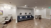 This single-floor salon and beauty spa is thoughtfully designed to deliver a polished, professional experience from the moment you enter. The space features a cohesive aesthetic defined by light wood-look flooring, crisp white walls, and recessed lighting that creates a bright, welcoming atmosphere throughout.

The styling area serves as a focal point, anchored by a professional salon chair positioned before a sleek white vanity and an illuminated mirror with elegant circular accent lighting. A dark feature wall adds dramatic contrast, while glass-paned double doors provide a refined entry point into this zone.

A dedicated nail care area offers pedicure stations alongside a built-in counter with white quartz surfaces and cabinetry. Vibrant macro floral artwork adds a lively, cheerful touch to the walls, contributing to a relaxing client environment.

A hair processing lounge features a tufted navy blue bench set within an alcove, complete with three overhead hooded dryers — ideal for coloring and conditioning treatments. Recessed lighting and neutral tones keep the atmosphere calm and comfortable.

A utility room provides dark wood cabinetry, a built-in sink with ample counter space, and generous upper storage — supporting the practical, behind-the-scenes needs of the salon with the same clean, modern finishes found throughout the rest of the space.