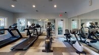 This single-floor fitness and amenity space is thoughtfully designed to offer a clean, functional environment for residents and users alike. The space is finished with a distinctive geometric patterned carpet in shades of gray, charcoal, and white, adding visual interest and a modern aesthetic throughout the common areas and exercise zones.

The fitness area is equipped with a selection of cardio machines, including treadmills, an elliptical, and a stationary bike, all positioned to take advantage of the natural light streaming through multiple windows. Full-length mirrors line one wall of the exercise area, enhancing the sense of openness and allowing users to monitor their form during workouts. A ceiling fan provides additional airflow and comfort. French doors offer direct access to an outdoor area, where seating is visible, seamlessly connecting the interior to the exterior environment.

The hallway and transitional areas feature the same cohesive flooring and neutral gray walls with white crown molding and baseboards, lending a polished, unified look across the space. Restroom facilities are conveniently accessible from the main corridor, along with louvered storage closets and a small built-in desk area. Recessed lighting throughout ensures consistent illumination, while HVAC vents and safety equipment such as fire extinguishers and exit signage reflect practical, code-compliant design. Overall, the space presents a bright, clean, and well-maintained environment.