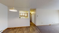 This single-floor unit offers a practical and well-organized layout designed for comfortable everyday living. The main living area features warm wood-look vinyl flooring and transitions seamlessly into a carpeted zone near the entry door, creating a natural separation of space. A pendant light fixture hangs overhead, adding a focal point to the open living and dining area. A distinctive pass-through window connects the living space to the kitchen, enhancing the sense of openness and allowing for easy interaction between rooms.

The galley-style kitchen is efficiently laid out with white cabinetry accented by warm wood trim, white appliances including a refrigerator, electric range, and dishwasher, as well as a stainless steel sink. Wood-look flooring continues throughout the kitchen, maintaining a cohesive aesthetic.

The bedrooms are carpeted and feature neutral walls, baseboard heating, and windows that allow natural light to filter in. One bedroom also includes a wall-mounted air conditioning unit for added climate control comfort.

The bathroom is clean and functional, featuring a full tub and shower combination with classic white tile surround, a single-sink vanity, and wood-look flooring. A hallway connects the main living areas to the bedrooms and bathroom, completing the straightforward and efficient floor plan.
