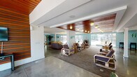 Located at 1718 Ximeno Ave in Long Beach, California, this single-story community space offers a sleek, modern lounge environment designed for comfort and social interaction. The open-plan layout is anchored by polished concrete floors that lend an industrial-chic aesthetic, while a striking warm wood-paneled ceiling adds richness and visual contrast to the bright white structural beams above.

The seating area is generously appointed with contemporary lounge chairs and modular sofas arranged to encourage both relaxed gathering and casual conversation. Soft overhead lighting fixtures complement the abundant natural light that pours in through expansive windows and glass openings along the perimeter.

A standout feature of the space is its seamless connection to an outdoor pool area, clearly visible through the large openings at the far end of the room. The aqua-toned accent walls echo the poolside palette, reinforcing the indoor-outdoor flow that defines the overall ambiance. Lush greenery adds organic warmth to the interior.

The overall atmosphere is bright, airy, and welcoming — a well-designed communal retreat that balances contemporary architectural details with practical, inviting furnishings. This space is well-suited for leisure, socializing, or simply enjoying the Southern California lifestyle from a comfortable indoor setting.