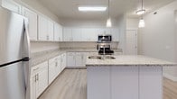 This single-floor residence presents a clean, contemporary aesthetic throughout its well-appointed layout. The kitchen serves as a central highlight, offering an abundance of white shaker-style cabinetry, light granite countertops, and stainless steel appliances including a range, microwave, and refrigerator. A kitchen island with a built-in sink provides additional prep space and a natural gathering point, while light wood-look flooring and overhead lighting complete the functional yet stylish design.

The living area is generously proportioned, featuring the same light wood-look flooring, track lighting, a ceiling fan, and a door that brings in natural light. The bedrooms are carpeted in neutral gray tones and receive ample natural light through large windows fitted with white horizontal blinds. The primary bedroom benefits from multiple windows and a ceiling fan.

Two full bathrooms mirror the kitchen's design sensibility, featuring white cabinetry, granite-topped vanities, large mirrors, and tub-shower combinations with light wood-look flooring. A dedicated laundry area houses a full-size top-loading washer and dryer alongside overhead shelving, adding everyday convenience to the space.